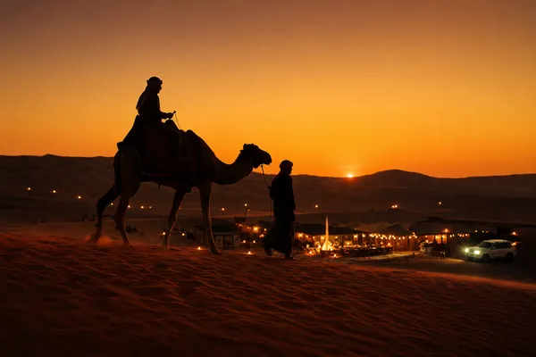 Camel trek on Lahbab red dunes at sunset above an evening desert safari camp